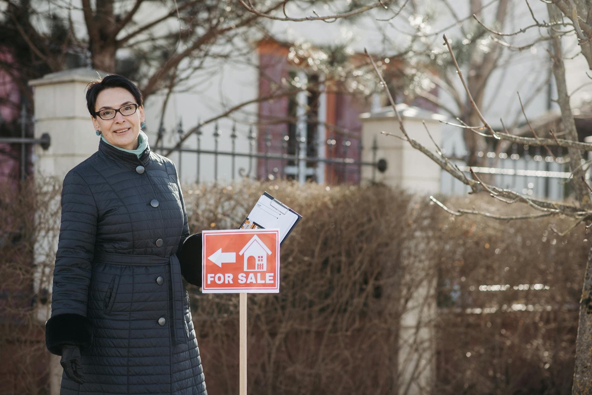 Smiling woman realtor in winter coat standing by a for sale sign with documents, outdoors.