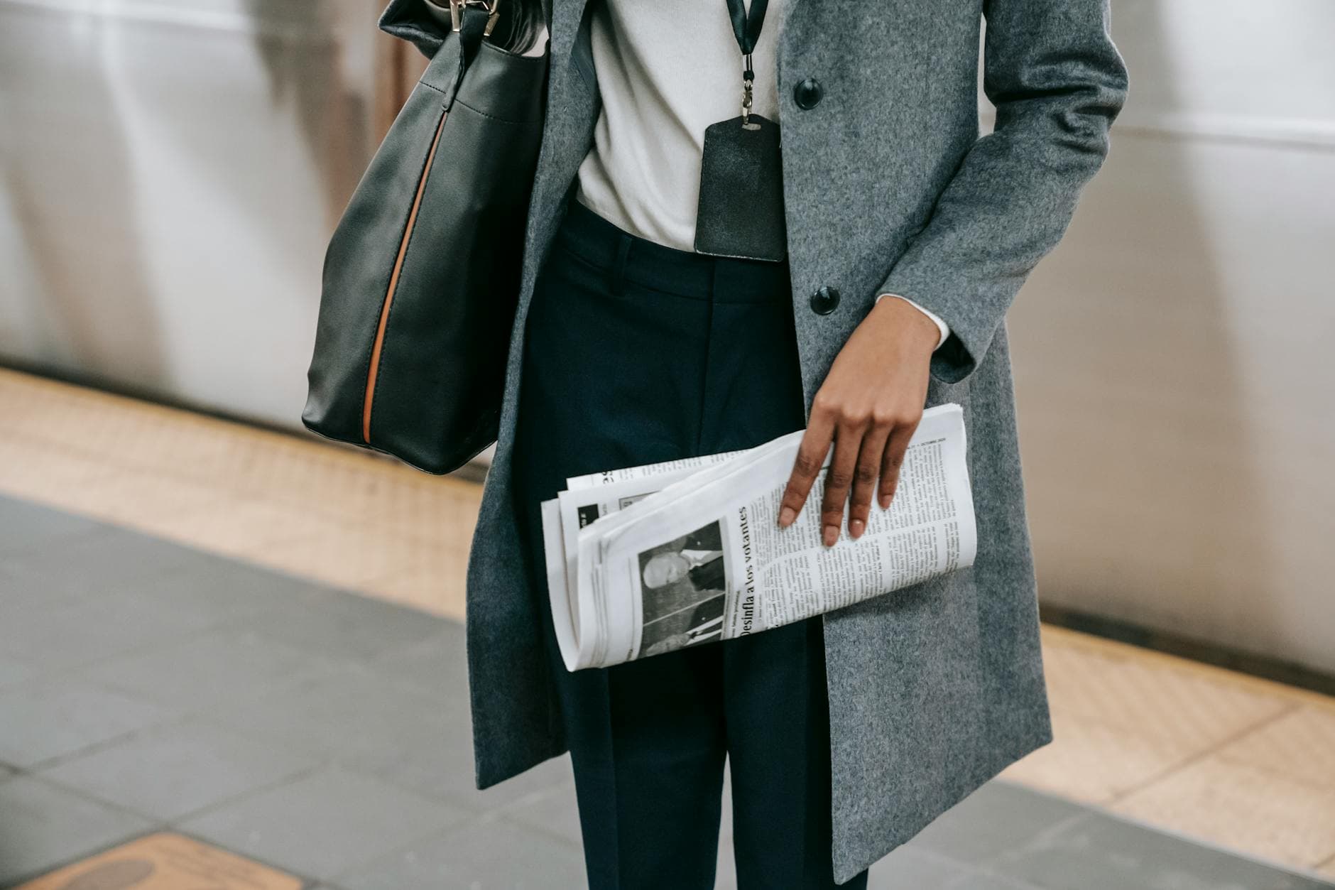 From above of crop anonymous female employee in elegant outfit and name tag standing on platform of metro station with newspaper in hand