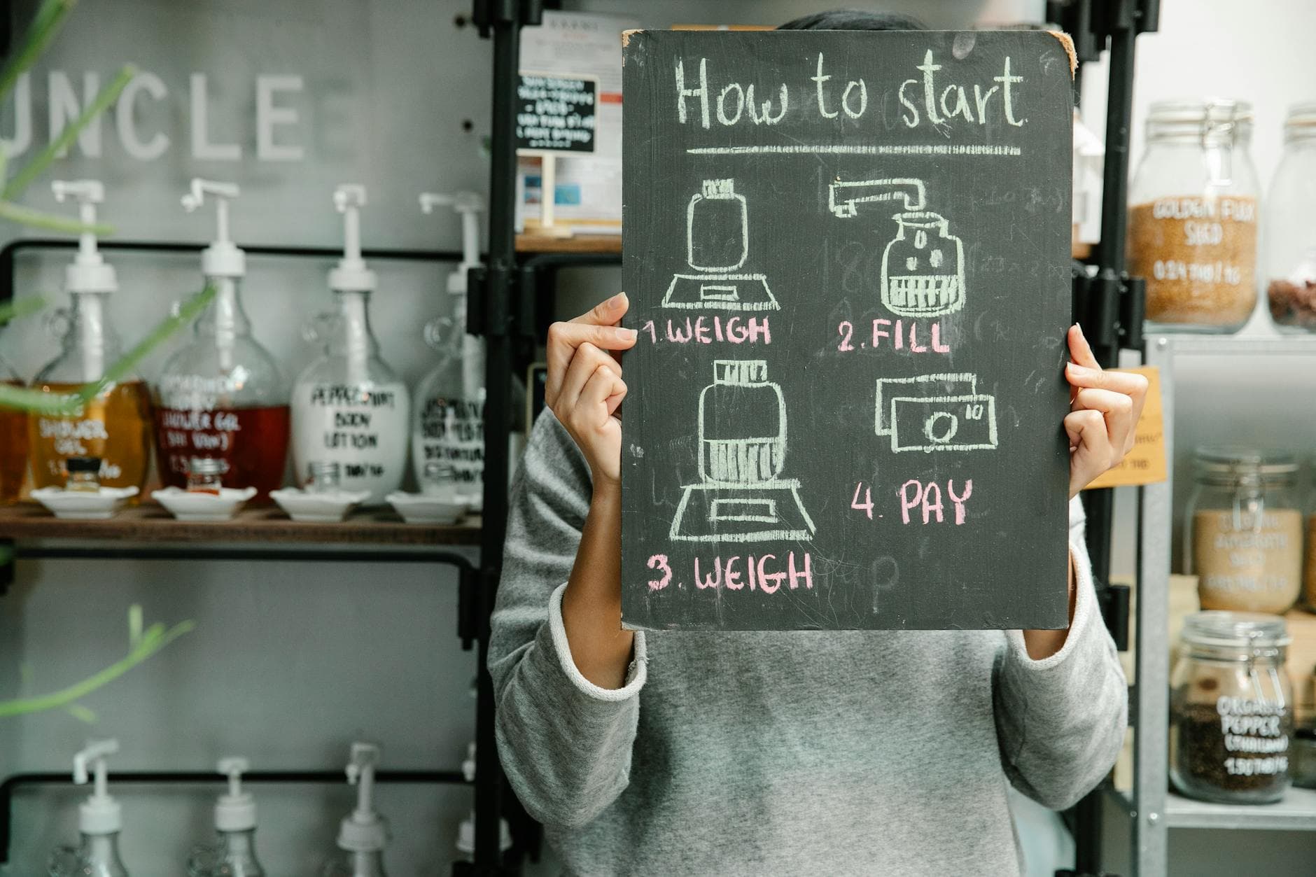 Person holding a chalkboard with shopping steps in a refill shop with glass containers on shelves.