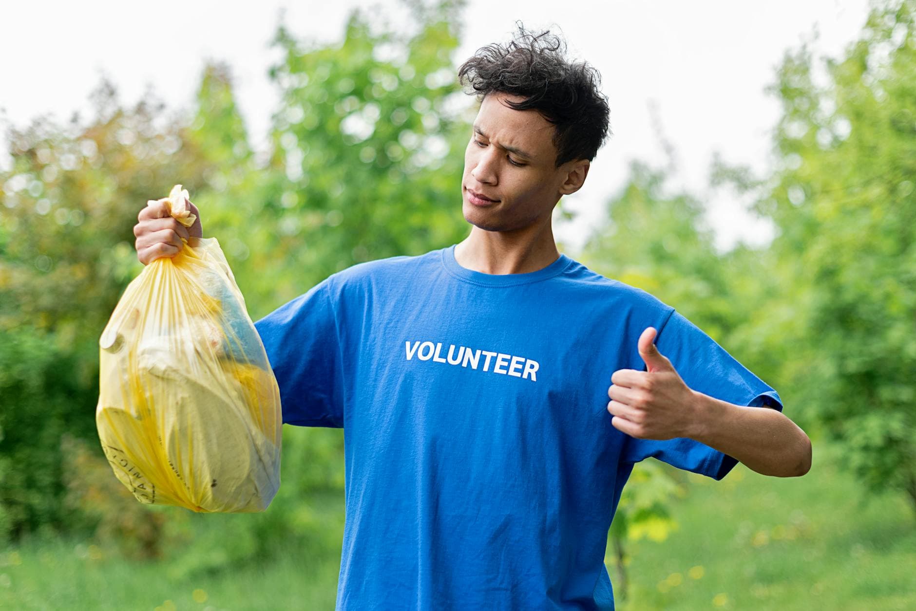 Young volunteer in a blue shirt holding a bag of recyclables and giving a thumbs up outdoors.