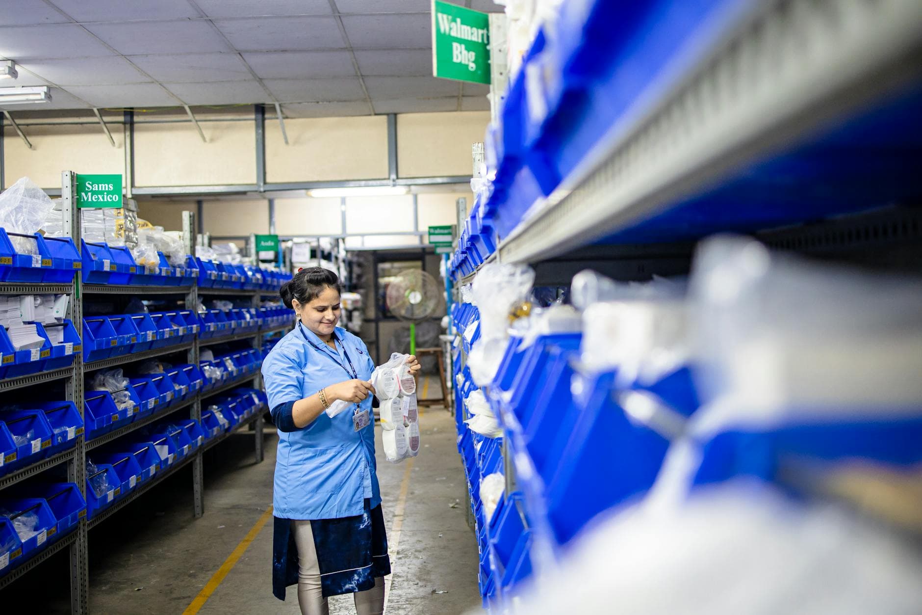 Female worker organizes shelves in a warehouse. Industrial setting with labeled storage bins.